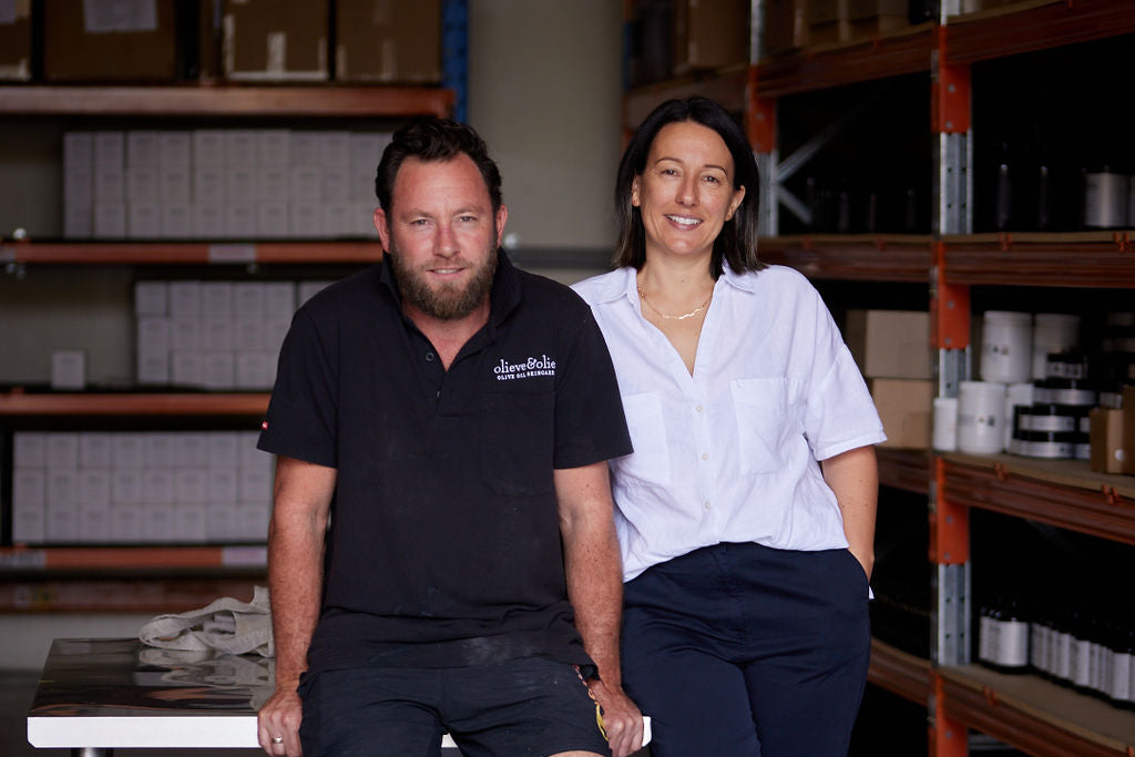 Sarah and Hal leaning against a workbench in the Rosebud factory, surrounded by shelves of finished Olieve & Olie products.