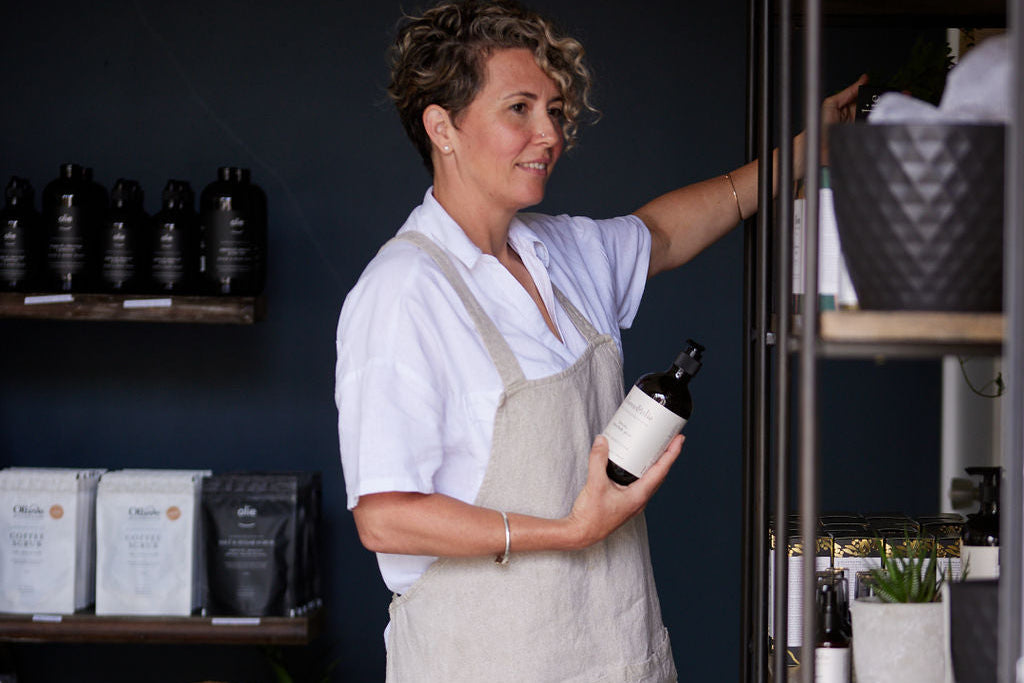 Tonika wearing a white shirt and beige apron holding an Olieve & Olie Hand & Body Wash in the factory shop, with shelves of products in the background.