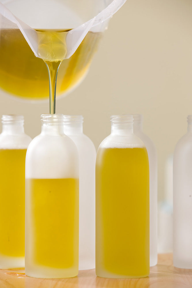 A close-up of Australian extra virgin olive oil being poured from a jug into frosted glass bottles on a wooden table, ready to be used in Olieve & Olie products.