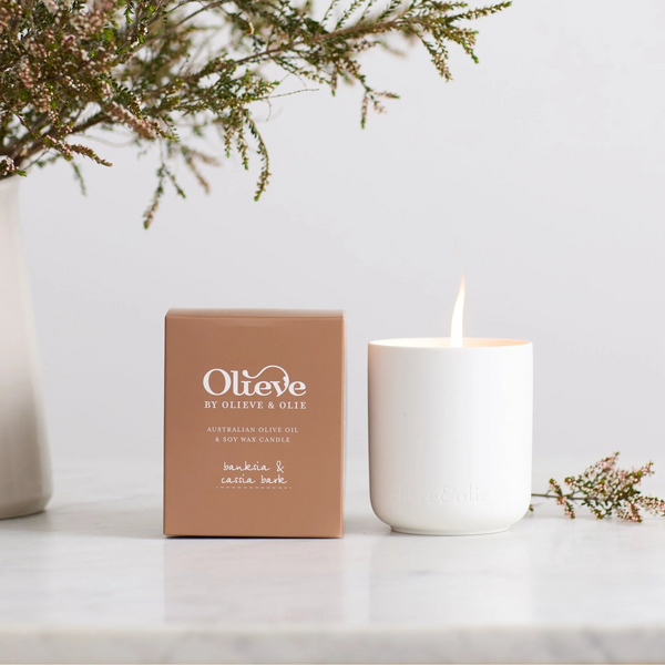 Olieve & Olie Banksia & Cassia Bark Candle displayed with its gift box on a marble bench, the candle softly lit beside a vase of fresh flowers, creating a warm and elegant atmosphere.