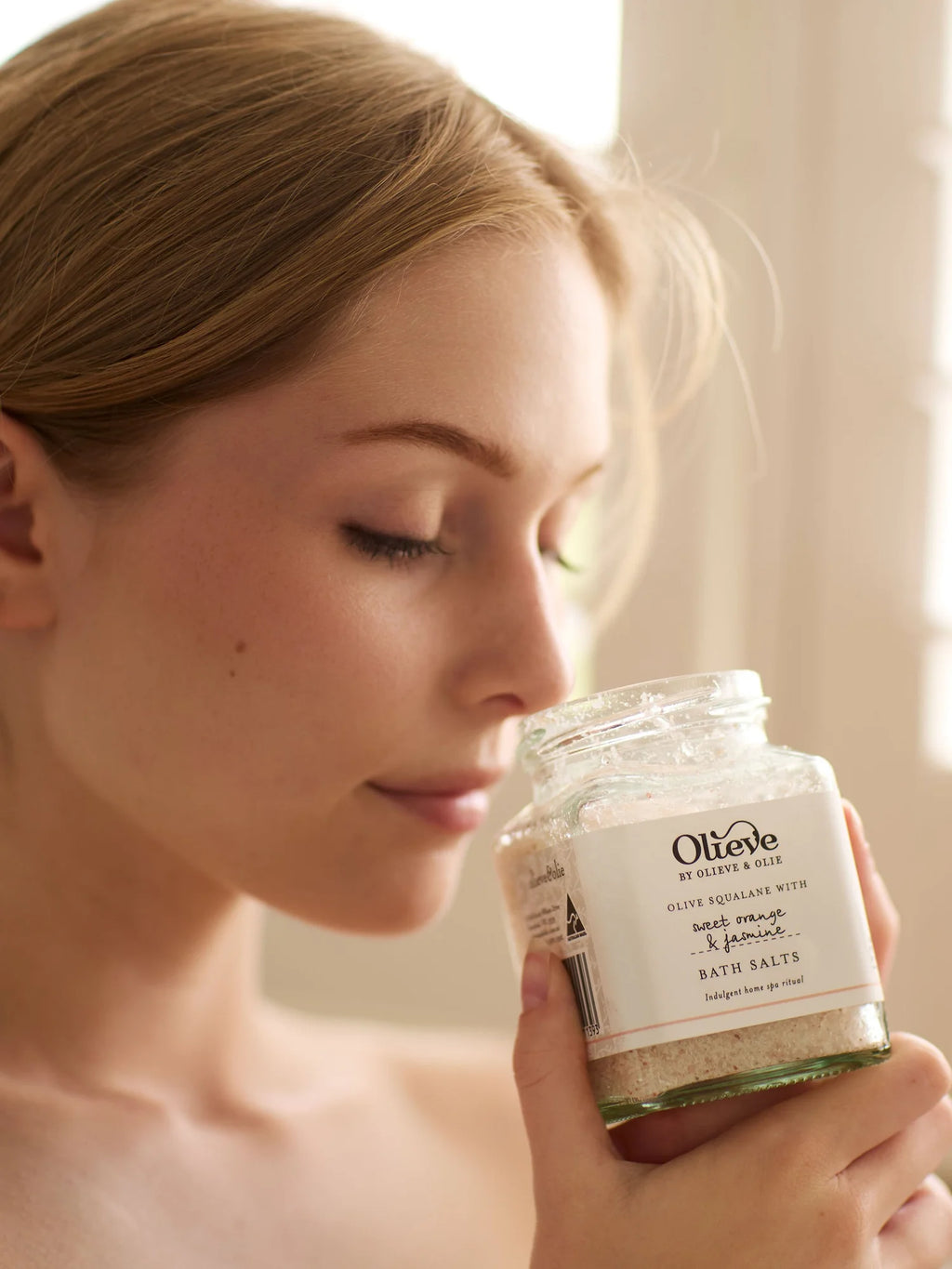 Woman enjoying the natural aroma of Olieve & Olie Bath Salts in a relaxing lifestyle setting. Made with a soothing blend of mineral-rich salts and botanical ingredients, this luxurious bath soak helps calm the senses, relax the body and soften the skin