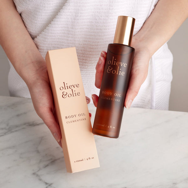 Hand holding an Olieve & Olie Clementine Body Oil above a marble bench. This lightweight botanical body oil, made with olive-derived squalane, deeply nourishes the skin while leaving it silky soft, smooth and naturally radiant