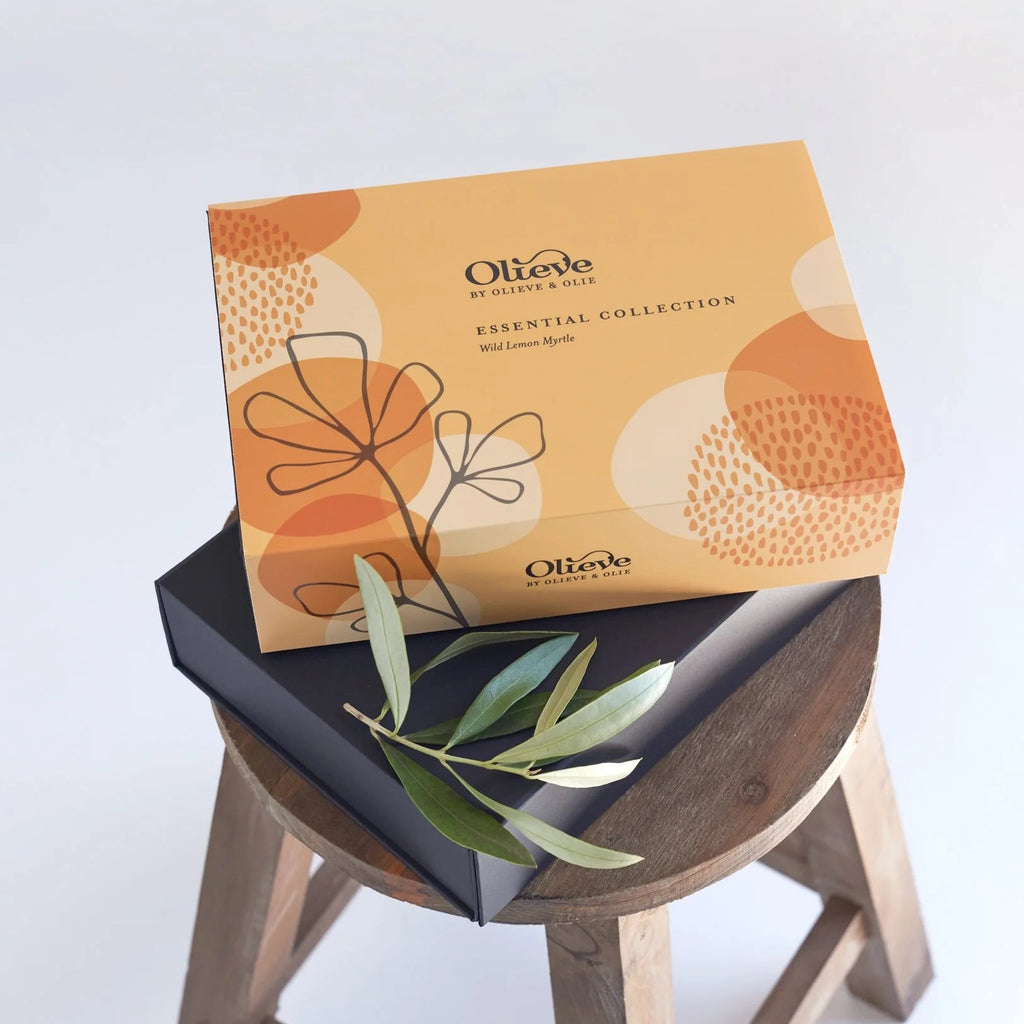 Olieve & Olie Essential Collection Lemon Myrtle Gift Set displayed on a wooden stool with fresh olive leaves. The beautifully presented gift box includes handcrafted natural skincare made with Australian olive oil, creating an elegant gift for any occasion