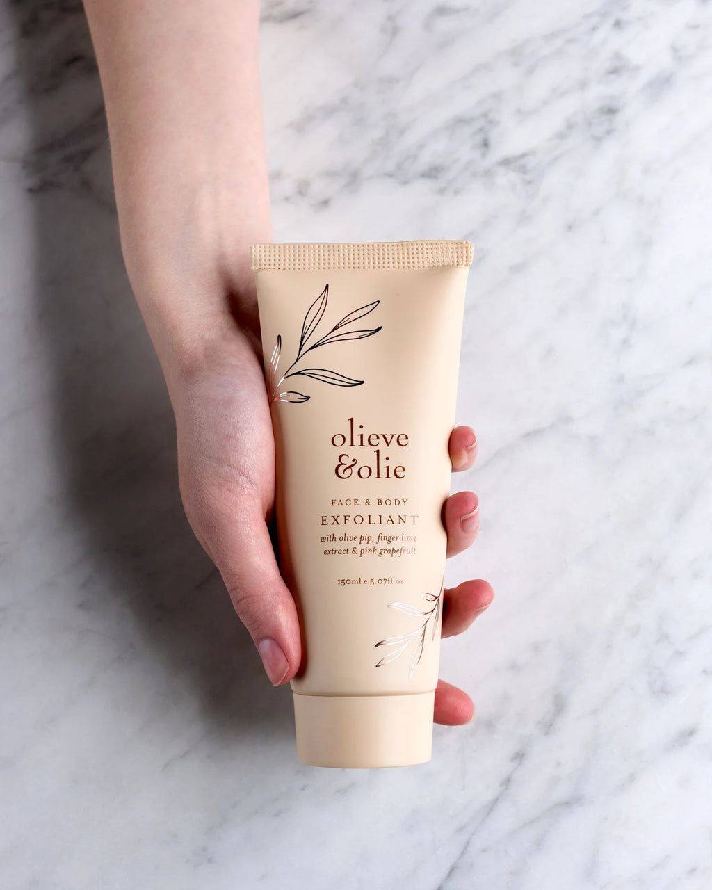 Hand holding an Olieve & Olie Face & Body Exfoliant above a marble table. This handcrafted exfoliating scrub gently buffs away dull skin with natural ingredients, leaving the face and body feeling smooth, soft and refreshed