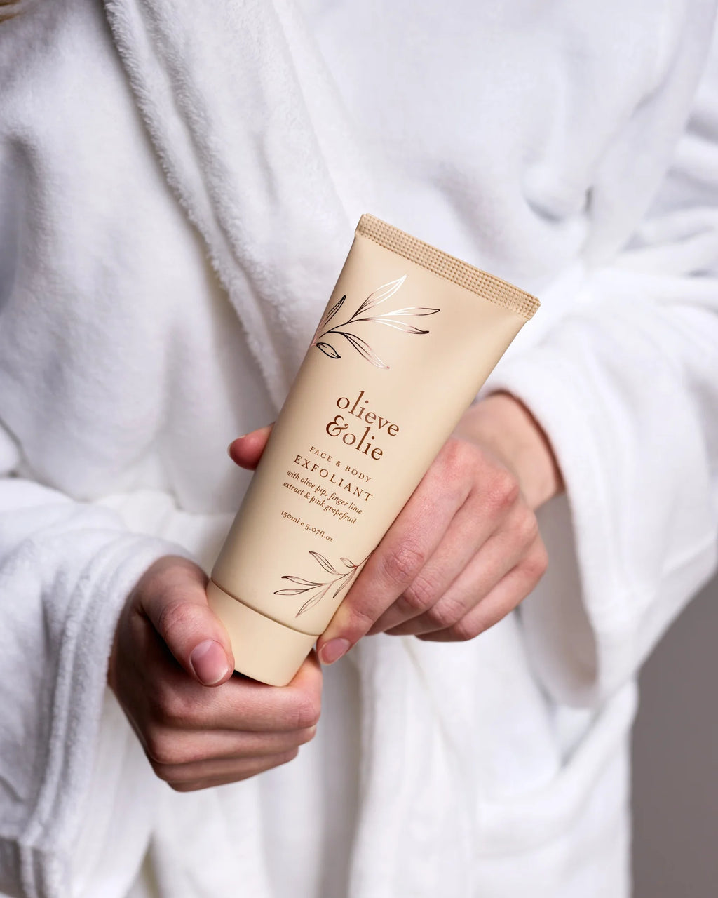 Person wearing a robe holding Olieve & Olie Face & Body Exfoliant in a relaxing spa-inspired setting. This gentle exfoliating scrub is handcrafted with natural ingredients to smooth, polish and refresh the skin, leaving it soft and radiant