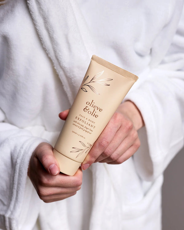 Person wearing a robe holding Olieve & Olie Face & Body Exfoliant in a relaxing spa-inspired setting. This gentle exfoliating scrub is handcrafted with natural ingredients to smooth, polish and refresh the skin, leaving it soft and radiant