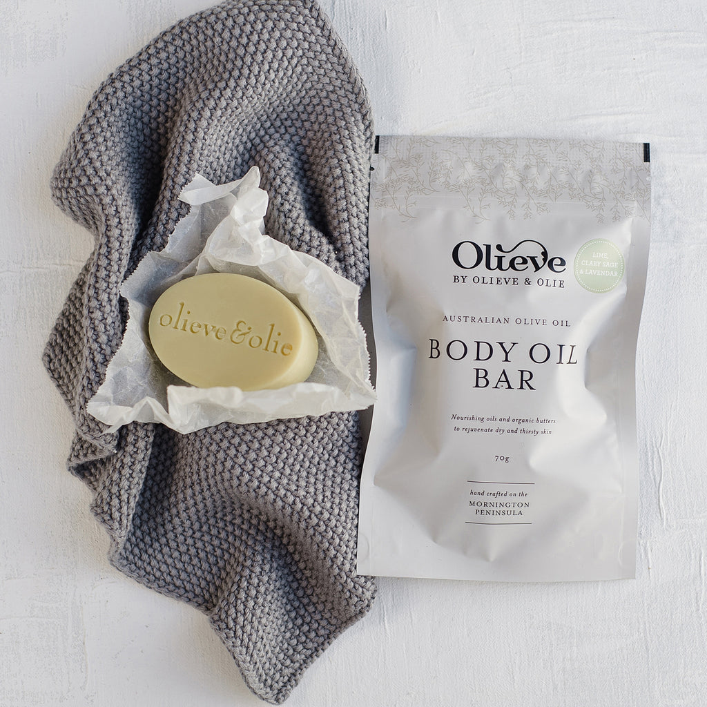 Olieve & Olie Lime, Clary Sage & Lavender Body Oil Bar displayed on a white background beside a grey knitted washcloth. The handcrafted solid moisturiser is made with Australian olive oil, cocoa butter and beeswax to deeply nourish and soften the skin