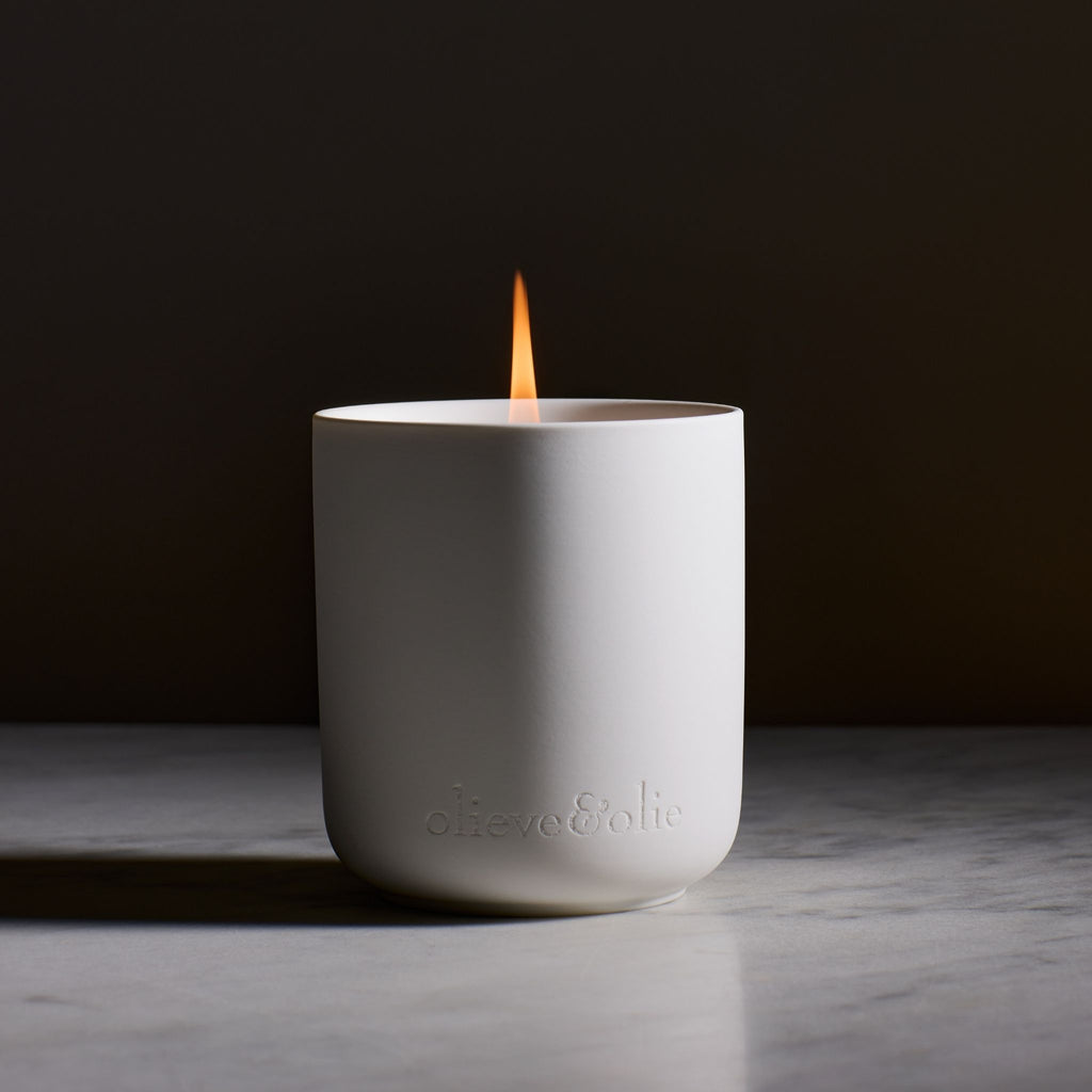 Olieve & Olie ceramic candle jar burning on a marble bench in a dark, moody setting. The softly glowing flame highlights the elegant white ceramic vessel, creating a warm and sophisticated home fragrance scene.