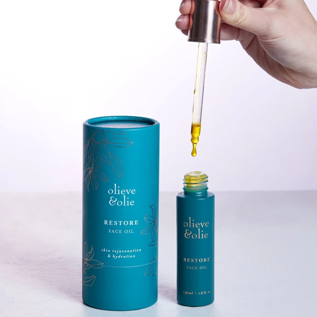 Person applying Olieve & Olie Restore Face Oil using the glass dropper. This nourishing facial oil, made with carefully selected botanical oils, helps deeply hydrate, replenish and restore the skin for a healthy, radiant complexion