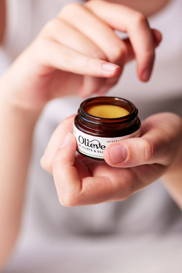 Hand holding an Olieve & Olie Vanilla Lip Balm in a natural lifestyle setting. Made with Australian olive oil and beeswax, this nourishing lip balm helps keep lips soft, smooth and deeply hydrated while showcasing its convenient everyday size