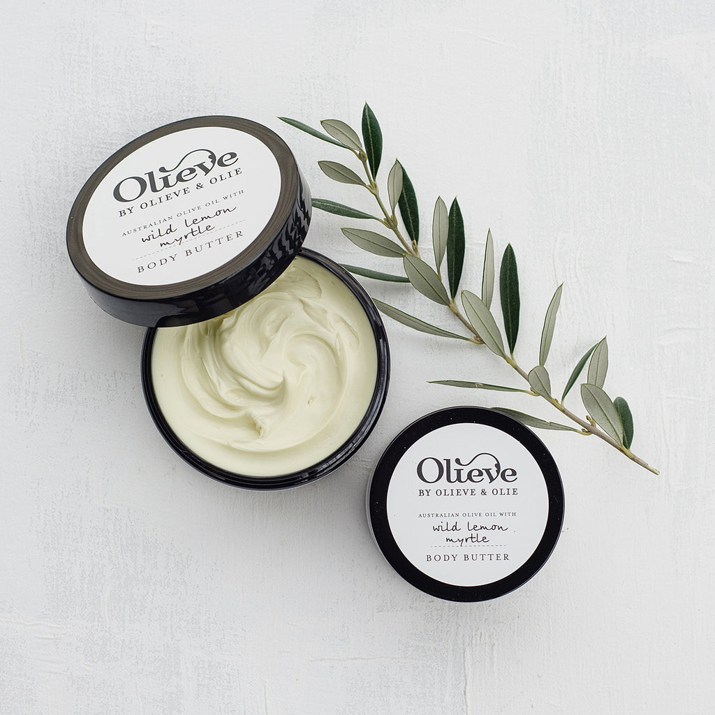 Open Olieve & Olie Wild Lemon Myrtle Body Butter with the lid beside the jar on a white background with a fresh olive branch. Made with Australian olive oil, cocoa butter and beeswax, this rich body butter deeply nourishes, softens and protects dry skin