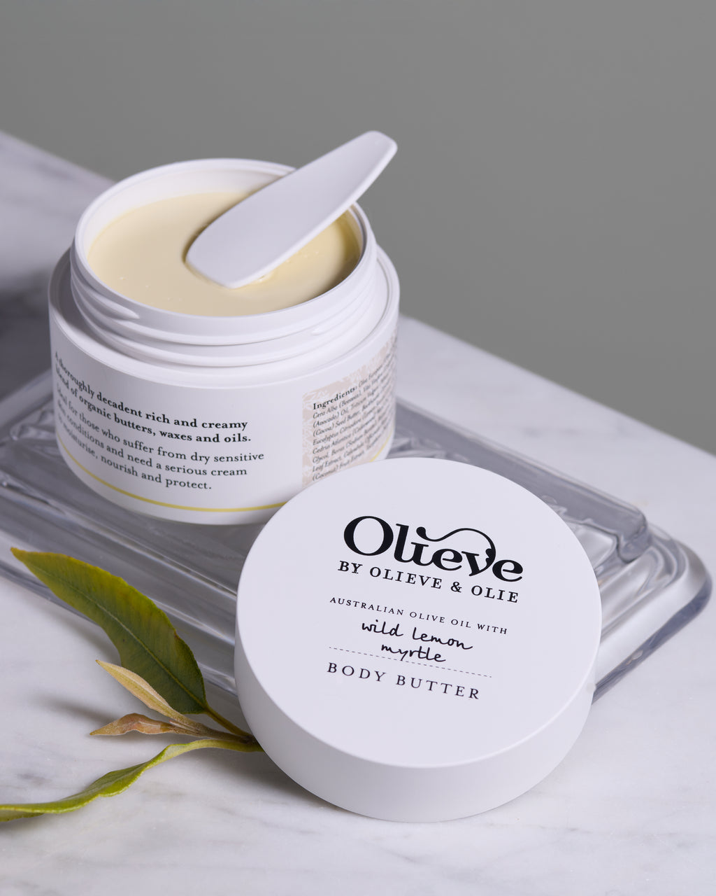 Open Olieve & Olie Wild Lemon Myrtle Body Butter displayed on a marble surface with the matching lid and fresh olive leaves. This rich, handcrafted moisturiser is made with Australian olive oil, cocoa butter and beeswax to deeply nourish and soften the skin