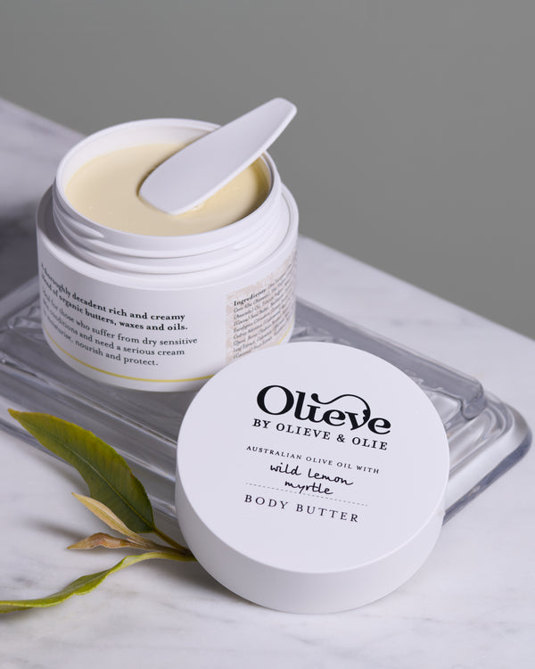 Open Olieve & Olie Wild Lemon Myrtle Body Butter displayed on a marble surface with the matching lid and fresh olive leaves. This rich, handcrafted moisturiser is made with Australian olive oil, cocoa butter and beeswax to deeply nourish and soften the skin