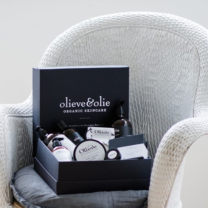 Olieve & Olie's skincare products in a matte black gift box sitting on a white lounge chair.